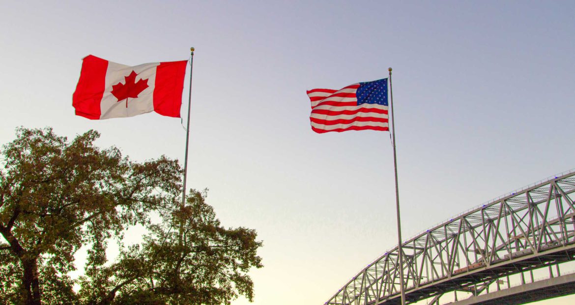 An American flag and a Canadian flag blow in the wind in the foreground. A tree grows in the background beside a tied arch bridge. The sun is setting and the sky is shades of bright yellows and deep blues.