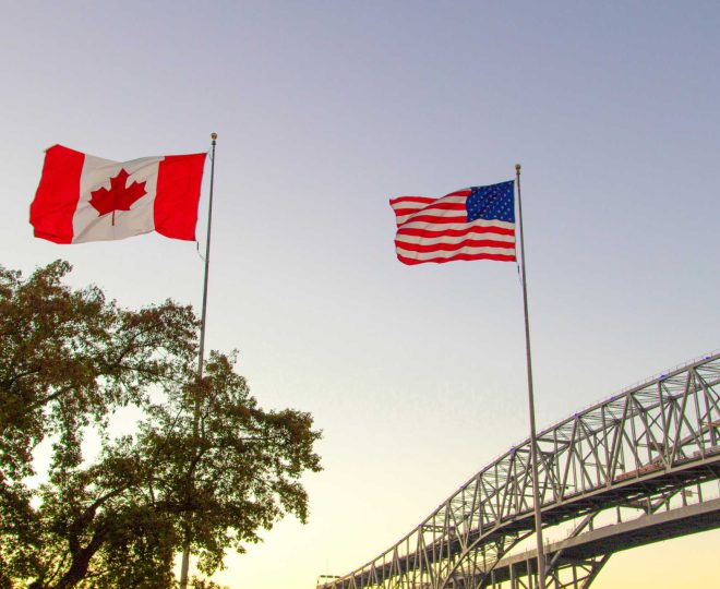An American flag and a Canadian flag blow in the wind in the foreground. A tree grows in the background beside a tied arch bridge. The sun is setting and the sky is shades of bright yellows and deep blues.