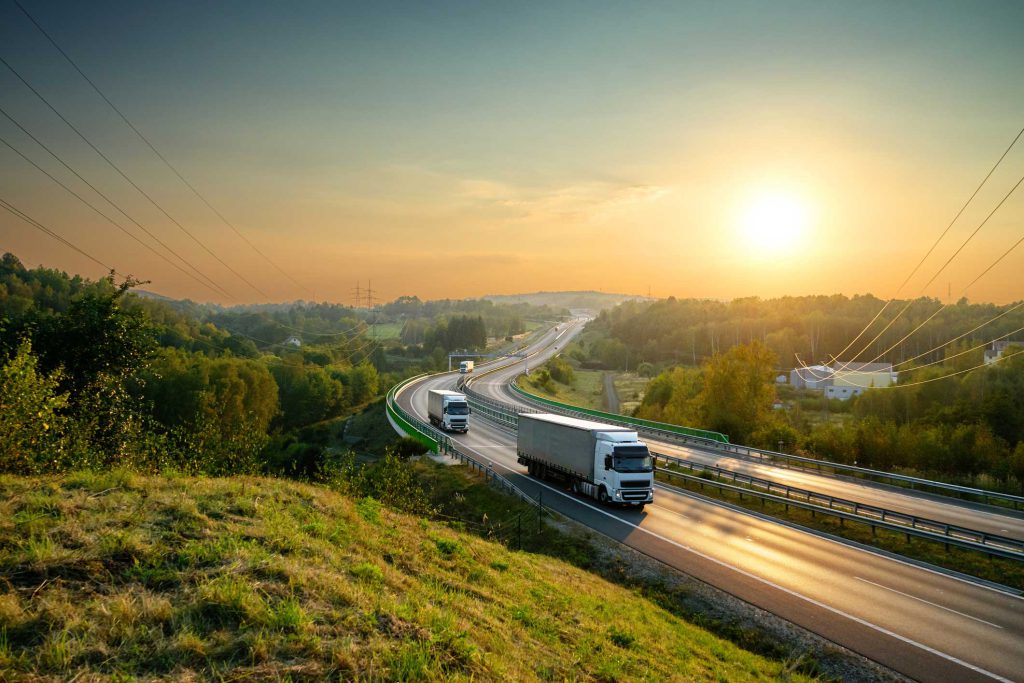 White trucks driving on the highway winding through forested landscape at sunset.