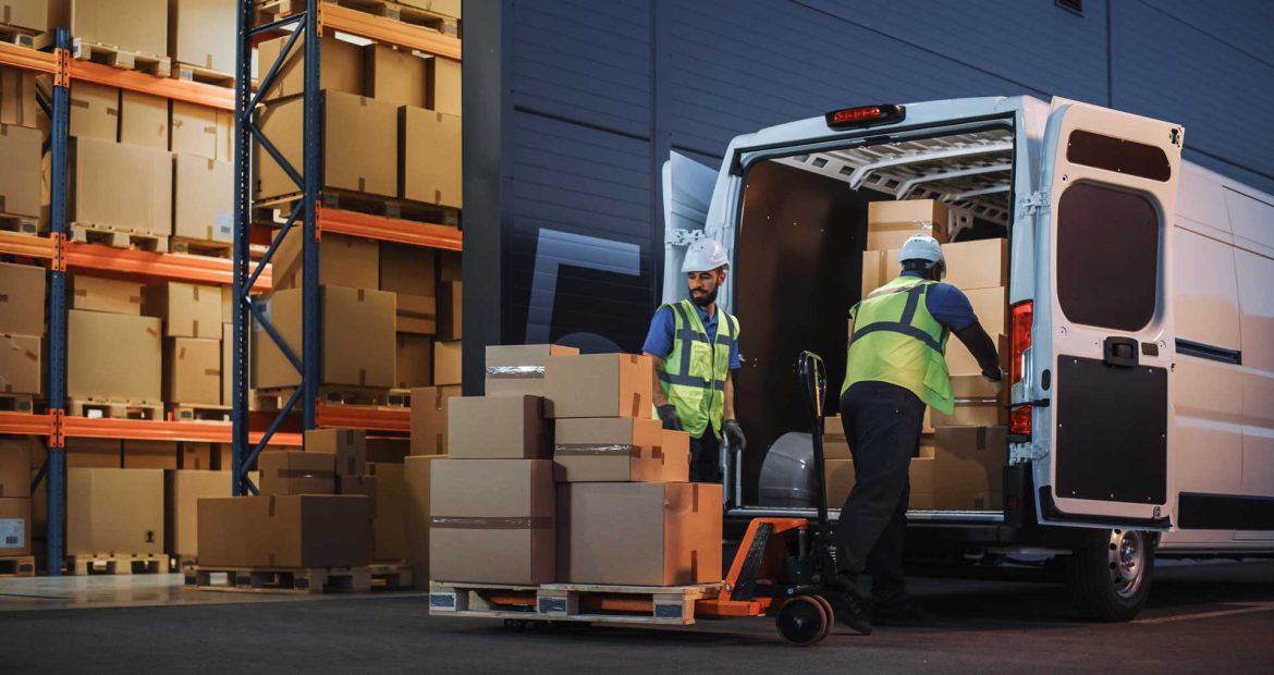 Two warehouse workers use a hand pallet truck to swiftly transfer goods between trucks at a cross-docking facility, minimizing storage and handling time in a busy industrial environment.